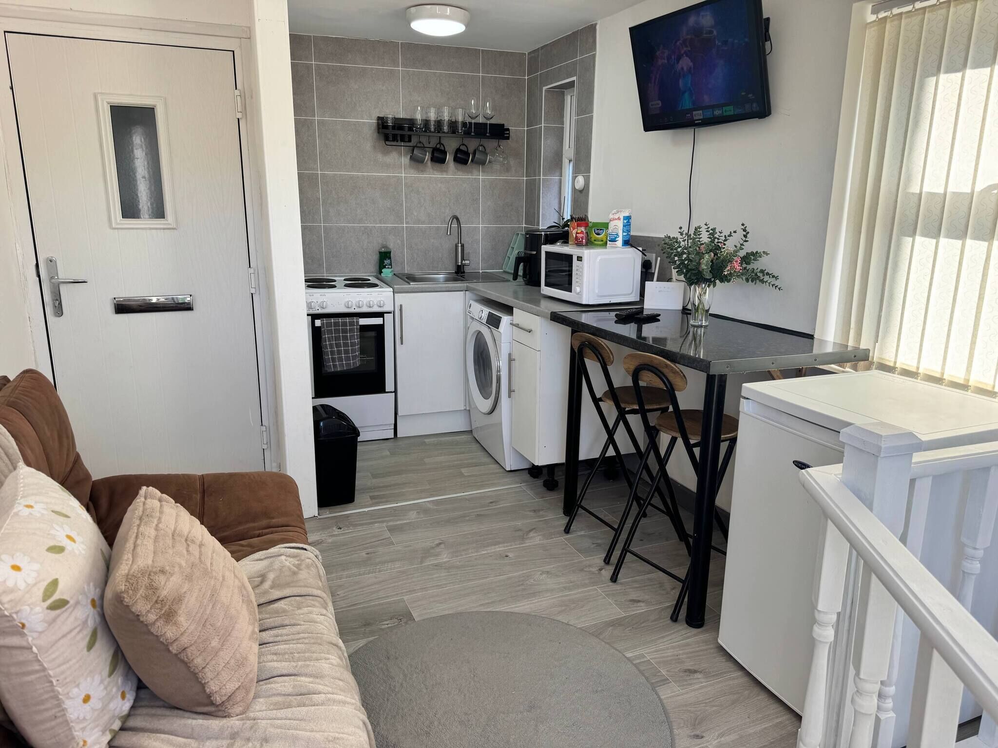 A compact kitchen with a microwave, stove, and washing machine, a dining area with chairs, and a TV mounted on the wall.