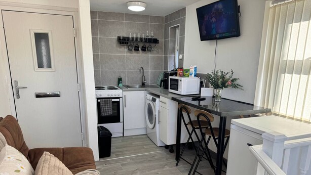 Uma cozinha compacta com micro-ondas, fogão e máquina de lavar roupa, uma área de refeições com cadeiras e uma TV na parede.