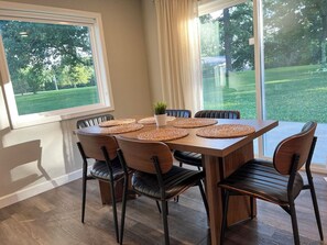 A dining area with a table set for six, surrounded by chairs, with a view of a green lawn through large windows.