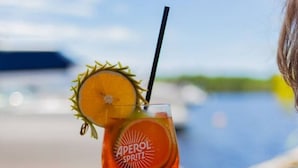 A cocktail glass with a straw, garnished with a slice of orange and a sprig, labeled 'APEROL SPRITZ' on a table by a poolside.