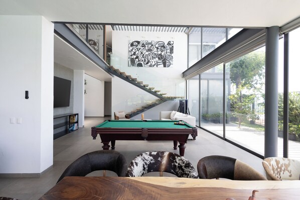 A modern interior with a pool table, a staircase, and a large window offering a view of the outdoors.
