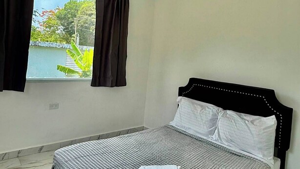 Un dormitorio con una cama, ventilador de techo, ventana con cortinas y vistas a zonas verdes.