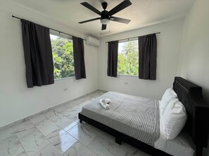 A bedroom with a bed, two windows with curtains, a ceiling fan, and an air conditioning unit. - The Rainforest Retreat! (Luquillo)
