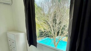 Una habitación con un aparador blanco, una ventana con cortinas negras y vistas a un árbol y una piscina exterior.