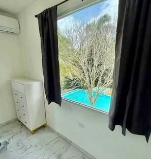 Ein Zimmer mit weißem Schminktisch, einem Fenster mit schwarzen Vorhängen und Blick auf einen Baum sowie einen Swimmingpool.