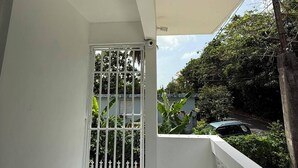 Un balcón con barandilla blanca, una puerta con reja metálica y vistas a zonas verdes y un coche aparcado.