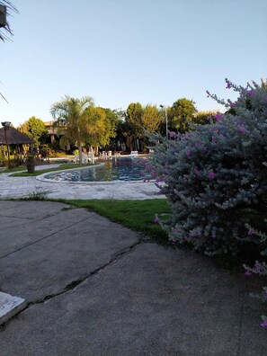 A pool area with a paved walkway, a thatched structure, and a bush with purple flowers. - Iloa Residence (Barra de São Miguel)