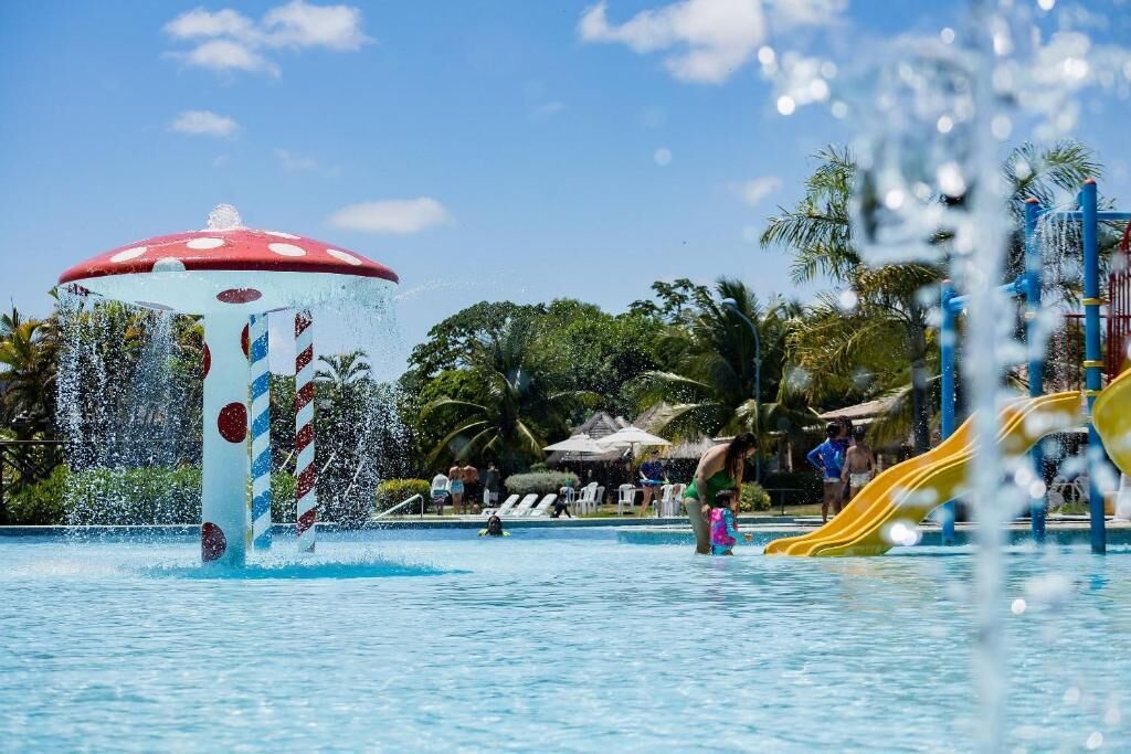 Un parco acquatico con una grande fontana a forma di fungo, uno scivolo giallo e persone che si divertono.