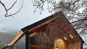 Uma cabana com fachada de madeira, grandes janelas, varanda e escadaria de madeira que leva à entrada.