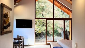 Um quarto espaçoso com teto alto, vigas de madeira, uma grande janela com vista para árvores, um sofá, uma TV fixada na parede e um espaço de jantar com mesa e cadeiras.
