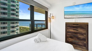 A hotel room with a large window overlooking a beach and a building, a bed with a rolled towel, a wooden dresser, and a wall-mounted TV. - Ocean View Large Condo in Beachfront Resort 1108 (Miami)