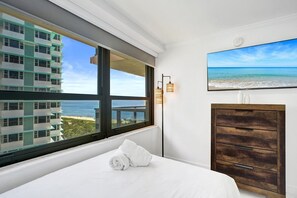 Ein Hotelzimmer mit großem Fenster mit Blick auf einen Strand und ein Gebäude, ein Bett mit zusammengelegtem Handtuch, eine Holzkommode und ein an der Wand befestigtes Fernsehgerät.