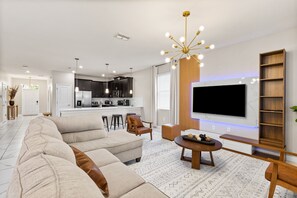 A modern living room with a large sectional sofa, a dining table, a TV mounted on the wall, and a kitchen in the background. - Themed 9BR Villa, Pool, Game Room, Champions Gate (Davenport)