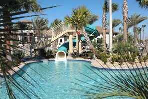 A swimming pool with a water slide, palm trees, and a building with a red roof in the background. - Themed 9BR Villa, Pool, Game Room, Champions Gate (Davenport)