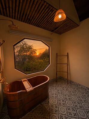 Een badkamer met een houten bad, een raam met uitzicht op een zonsondergang en geplakte vloertegels.