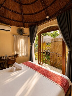 Een kamer met een bed, een tafel, een stoel, een ventilator en uitzicht op een tuin via een glazen deur.
