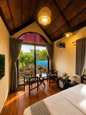 A room with a bed, a table with chairs, a ceiling fan, and a view of greenery outside. - Sora Tam Coc  (Nam Hoa Lu)