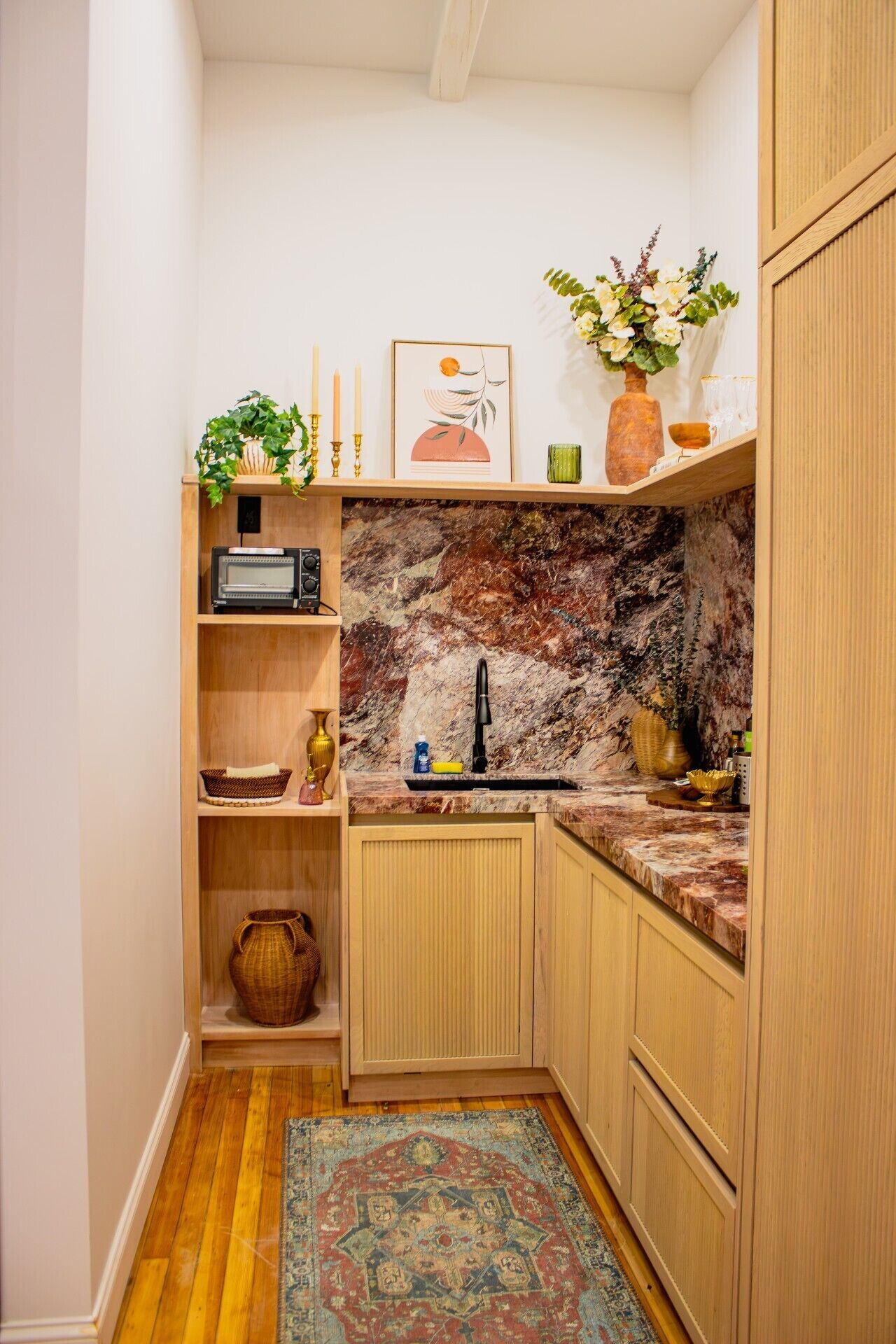 Uma cozinha compacta com painel de fundo de mármore, armários de madeira, uma pia e uma pequena prateleira com objetos decorativos.