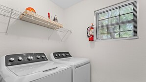 Un espace buanderie comprenant deux machines à laver à chargement frontal, une étagère avec des produits de lessive et un extincteur fixé au mur.