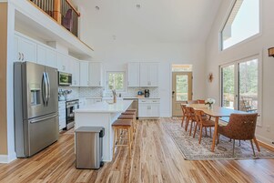Una cocina moderna con armarios blancos, electrodomésticos de acero inoxidable y una zona de comedor con sillas y una mesa de madera.