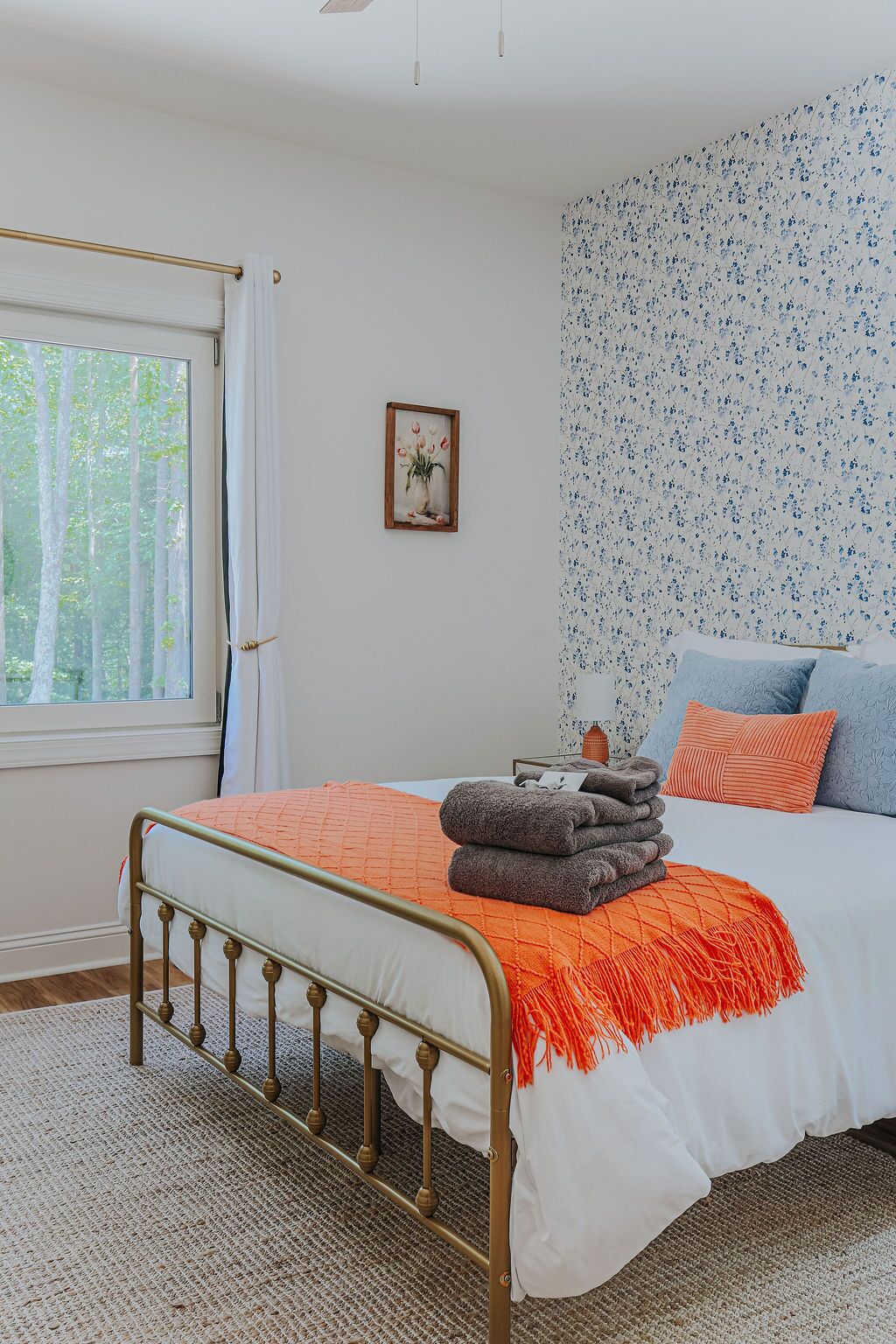 A bedroom with a bed, a window, a wall with floral wallpaper, and a framed picture on the wall.