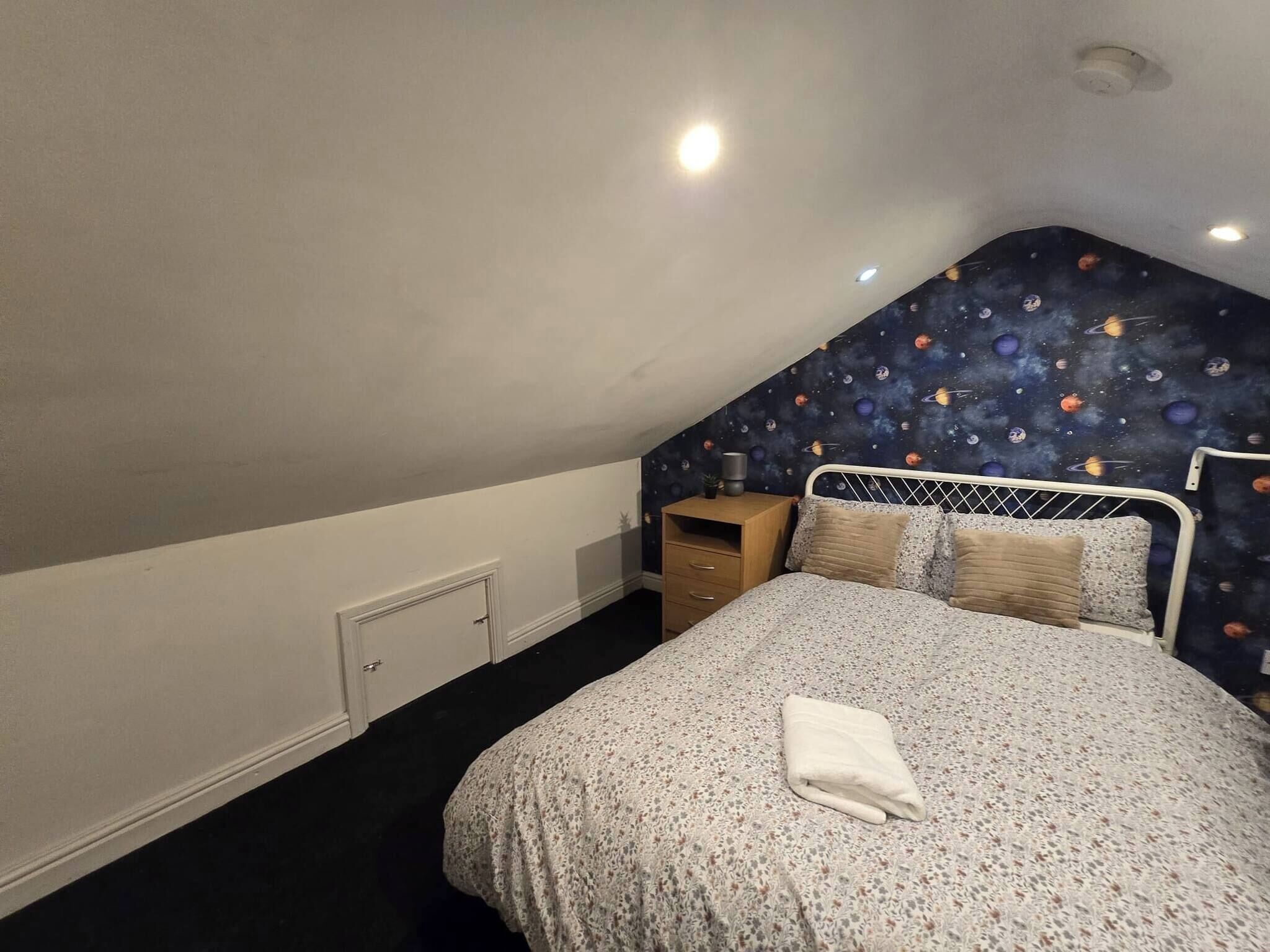 Um quarto com teto inclinado, uma cama com colcha estampada, uma mesa de cabeceira com abajur e uma parede com um mural com tema espacial.