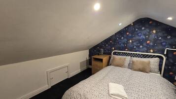 Un dormitorio con techo inclinado, una cama con colcha estampada, una mesita de noche con lámpara y una pared con un mural de temática espacial.