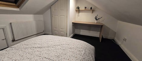 Un dormitorio con cama, un radiador, una puerta, una estantería con plantas y un escritorio.