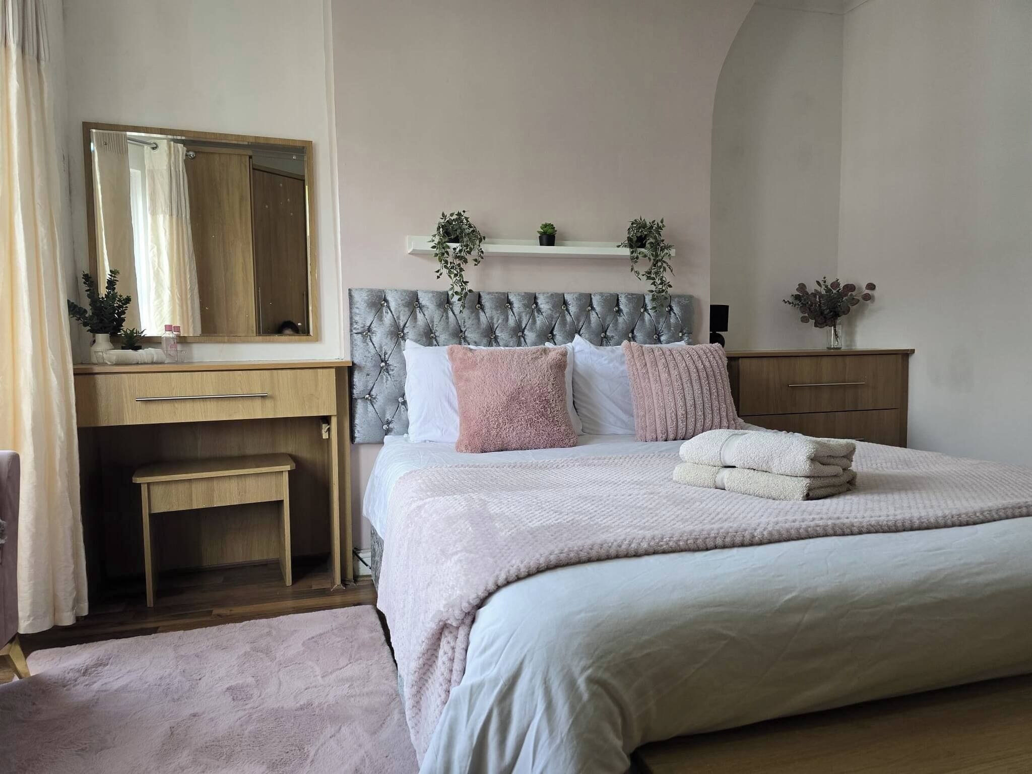 Quarto com cama, mesa de cabeceira, espelho, penteadeira e um pequeno banco.