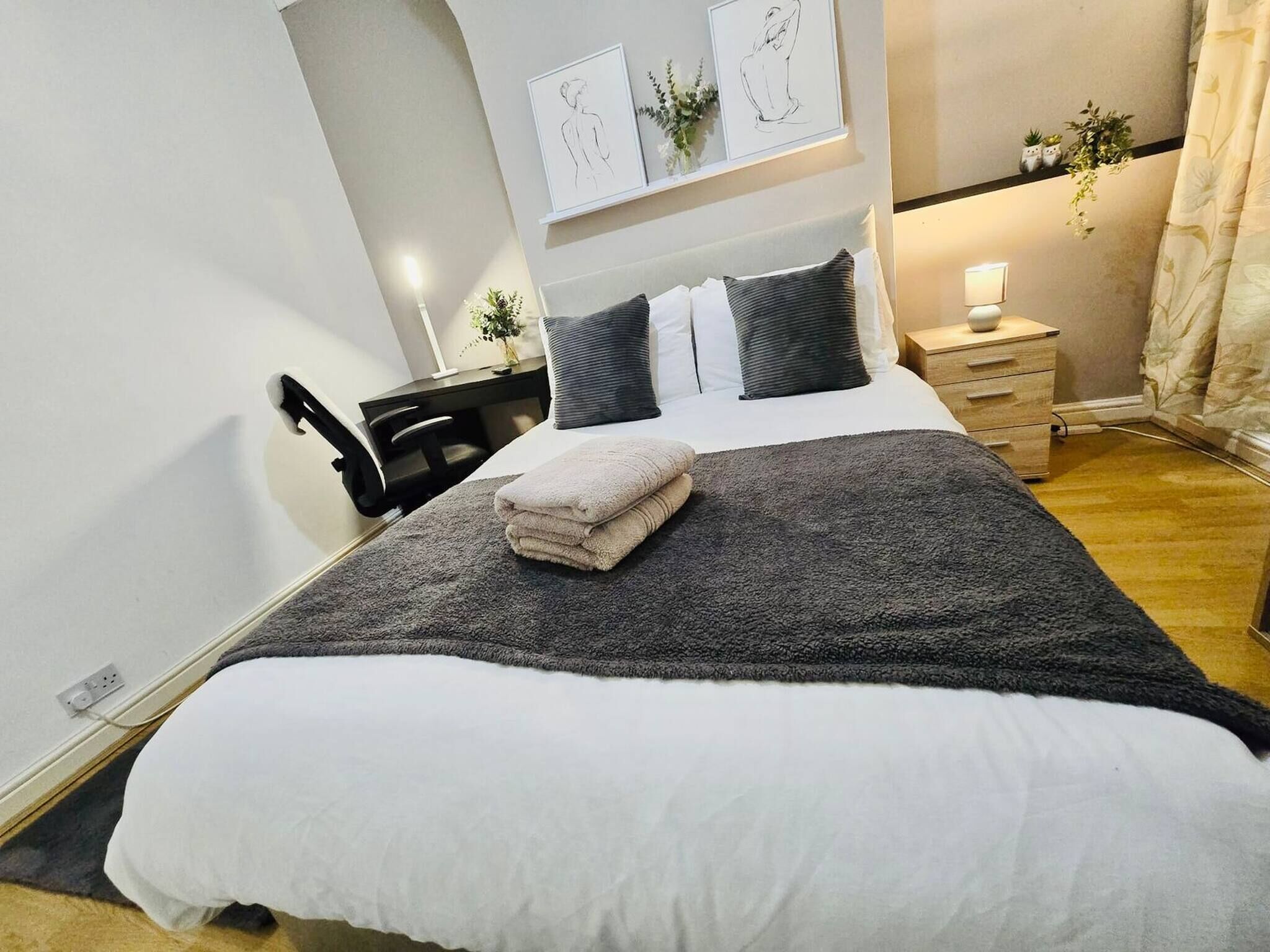 A neatly made bed with a grey blanket and two pillows, a bedside table with a lamp, and a small plant on the nightstand.
