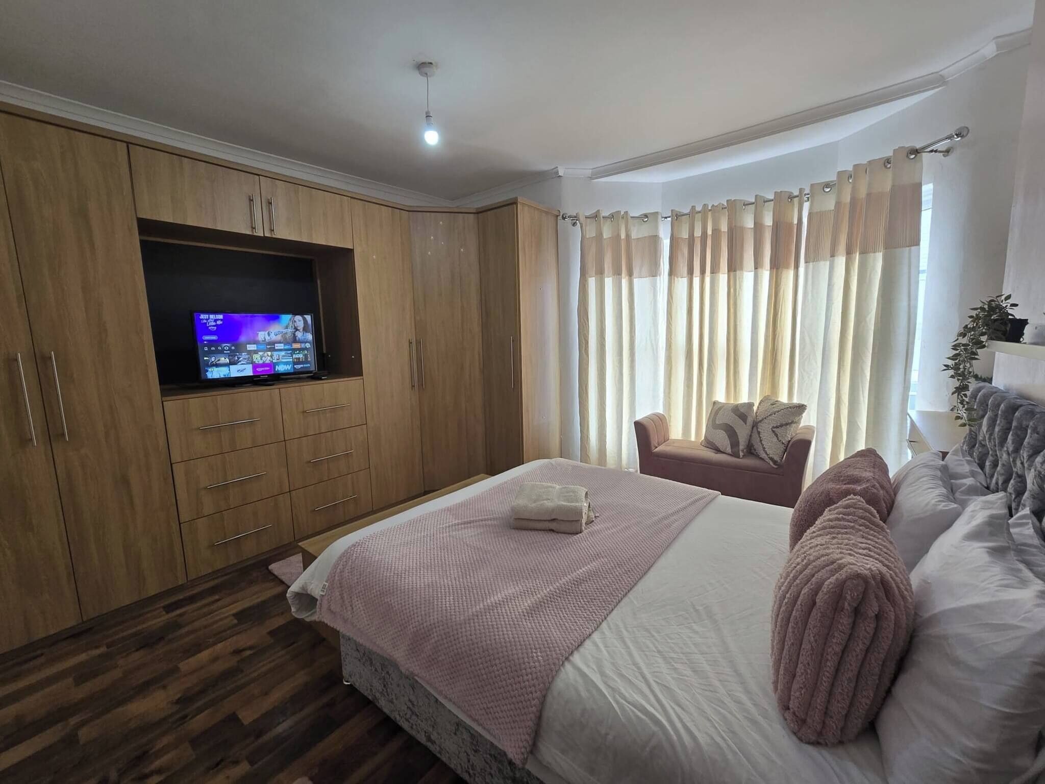 Ein Schlafzimmer mit einem Bett, einem an einer Holzeinrichtung montierten Fernseher, einer Couch und einem Fenster mit Vorhängen.