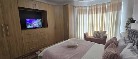 Ein Schlafzimmer mit einem Bett, einem an einer Holzeinrichtung montierten Fernseher, einer Couch und einem Fenster mit Vorhängen.