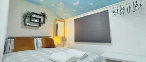 Un dormitorio con cama, mesita de noche, estante en la pared con plantas, ventana con persianas y lámpara en el techo.