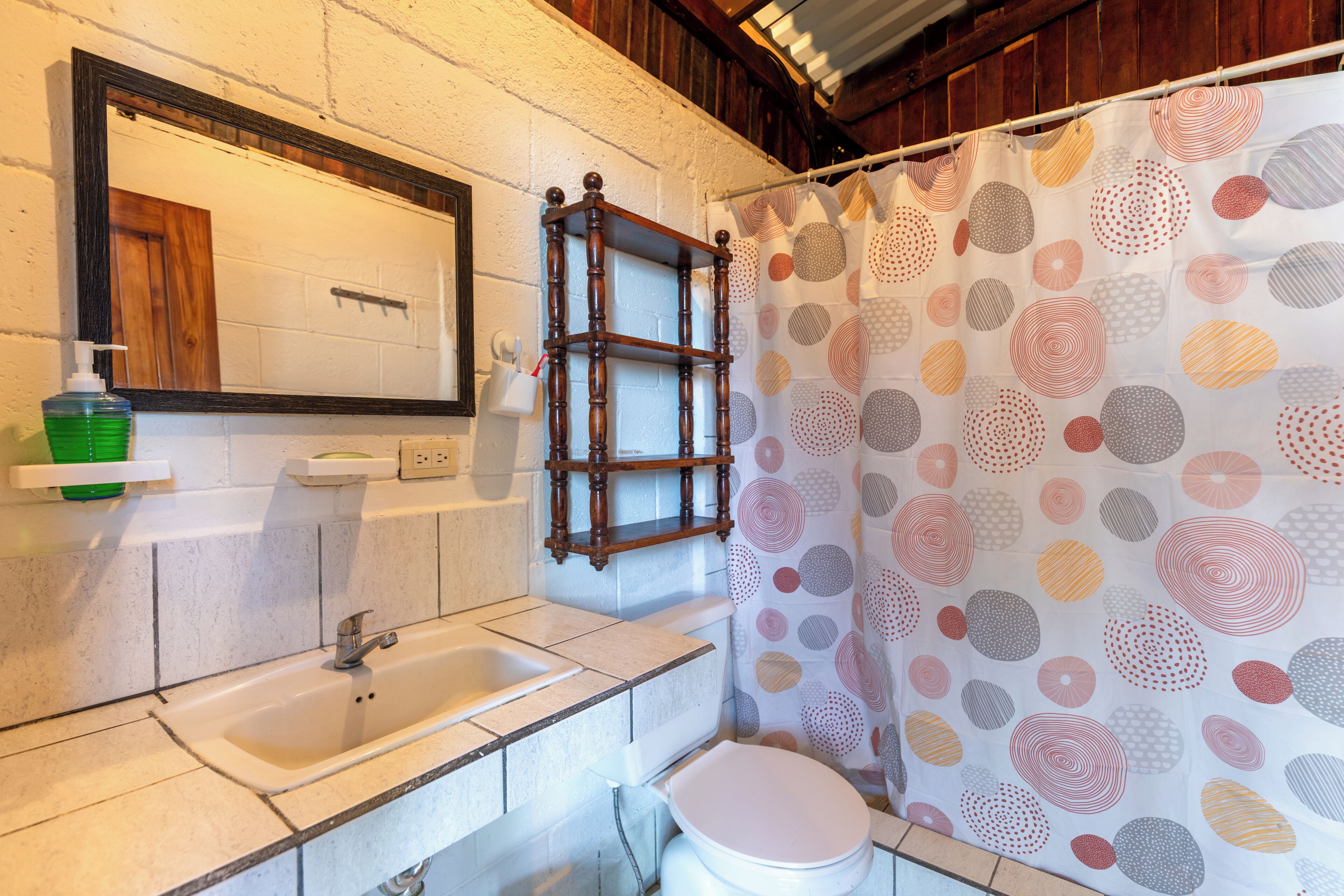 A bathroom with a sink, mirror, and toilet, a wooden shelf, and a patterned shower curtain.