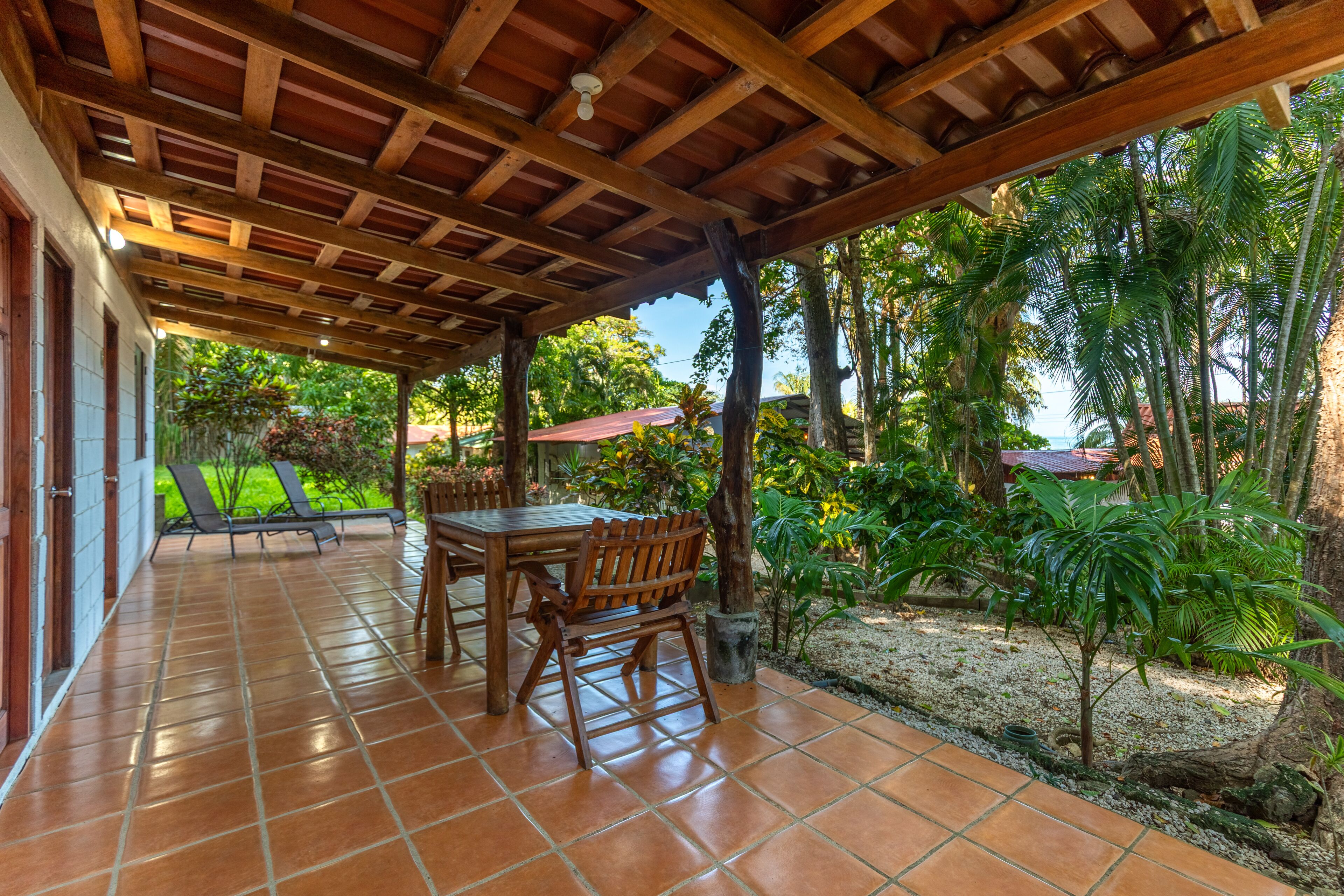 A covered patio with a tiled floor, wooden furniture, and a view of a lush garden and palm trees.