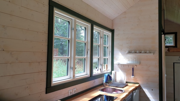A wooden cabin interior with a large window, a sink, and a shelf with cups. - A cozy little house in the countryside (Nutteln)