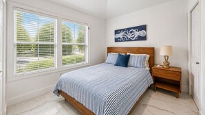 Ein Schlafzimmer mit einem Bett, einem Nachttisch, einem Deckenventilator, einem Fenster mit Jalousien und einem Wandbild.