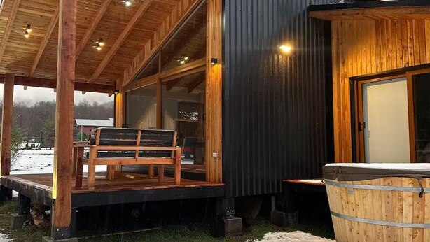 Eine Holzhütte mit überdachter Veranda, Whirlpool und Blick auf eine verschneite Landschaft.