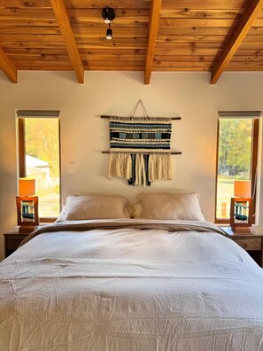 A bedroom with a large bed, two bedside lamps, a wall hanging, and windows with blinds.