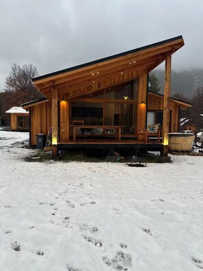 A wooden cabin with a large glass window, a deck, and a snow-covered ground.