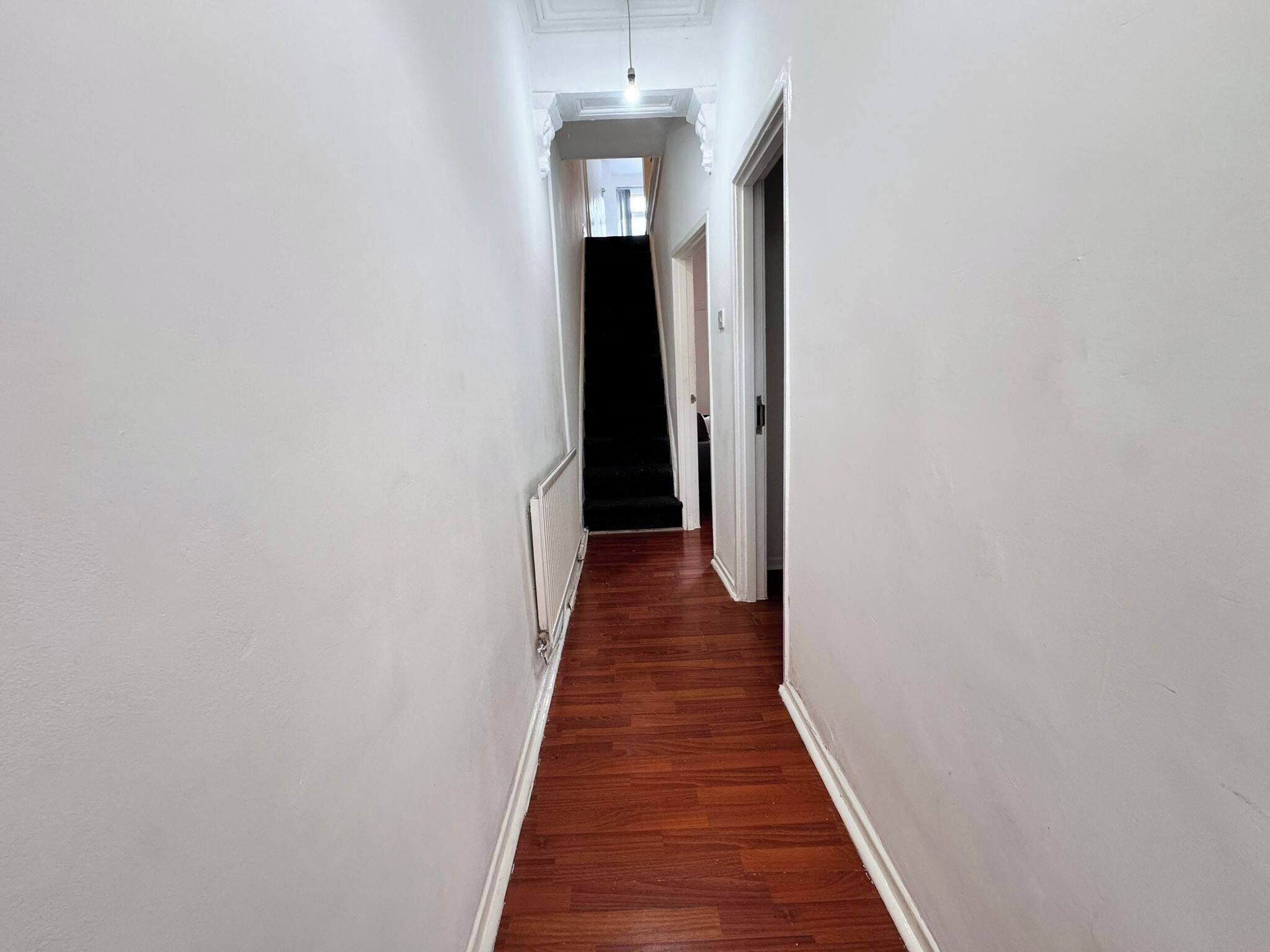 Passadís estret amb parets blanques i terres de fusta, que porta a una habitació fosca.