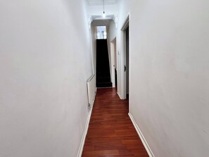 Un pasillo estrecho con paredes blancas y suelo de madera, que conduce a una habitación oscura.
