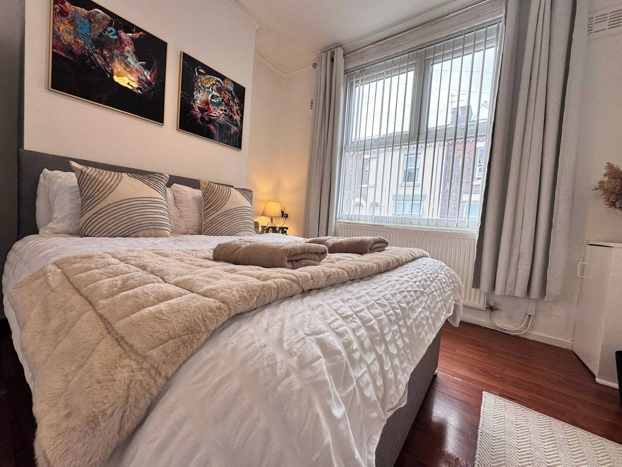 Una cama bien hecha con colcha y dos toallas, una ventana con persianas, y dos cuadros en la pared.