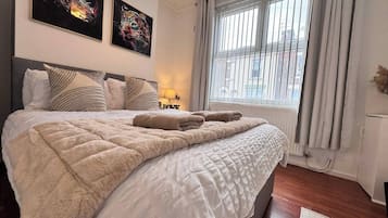 A neatly made bed with a quilt and two towels, a window with blinds, and two framed pictures on the wall.