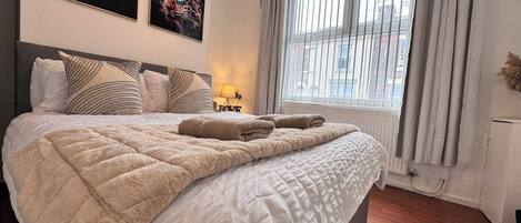 A neatly made bed with a quilt and two towels, a window with blinds, and two framed pictures on the wall.