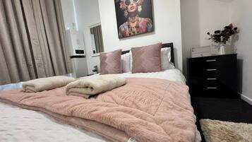 Een opgemaakt bed met een roze sprei, een schilderij aan de muur en een zwarte dressoir met lades.