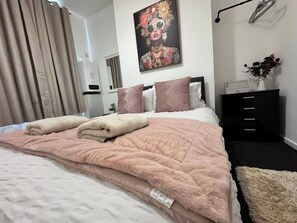 Ein ordentlich bezogenes Bett mit einer rosa Quilttdecke, ein Bild an der Wand und eine schwarze Kommode mit Schubladen.