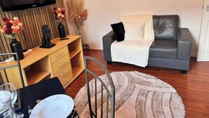 A living room with a grey sofa, a wooden TV stand, a dining table with chairs, and a large flat-screen TV. - House 10 Minutes From the Center (Liverpool)