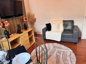 Una sala de estar con un sofá gris, un mueble bajo de madera para la televisión, una mesa de comedor con sillas y un televisor de pantalla plana grande.