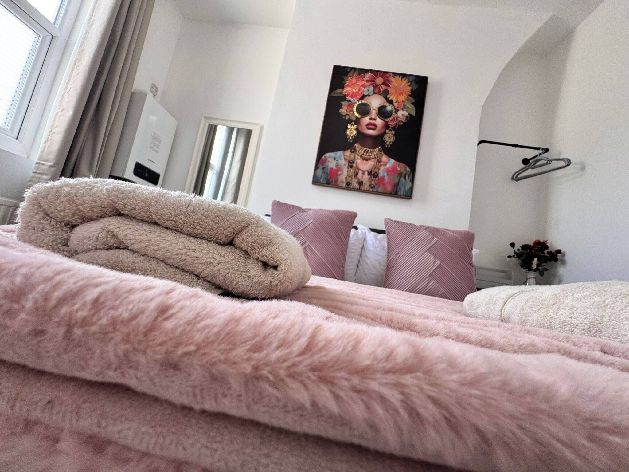 Uma cama bem arrumada com um cobertor rosa fofo, um quadro com um arranjo floral na parede e uma janela com cortinas.